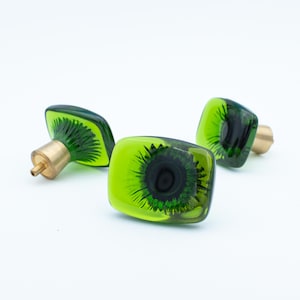 May include: Three decorative cabinet knobs with a unique kiwi fruit design. The knobs feature a square shape with a translucent green exterior and a dark center, complemented by brass hardware.