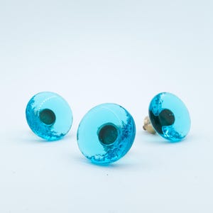 Puede incluir: Tres pomos circulares para armarios con una cúpula de resina azul turquesa brillante y translúcida con un patrón interior orgánico y texturizado, y una base de montaje metálica visible en la parte posterior de cada pieza, dispuestos en grupo.