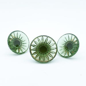May include: Three circular buttons made of translucent sage green glass. Each button features a central circular indentation surrounded by a radiating sunburst pattern of etched lines that extend toward the smooth, rounded outer edge.