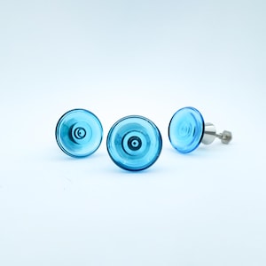 May include: Three blue glass stud earrings with a circular design. The earrings have a transparent, layered appearance with a silver post and backing. The earrings are set against a white background.