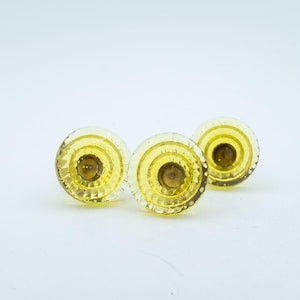 Puede incluir: Tres pomos circulares de cristal amarillo con un diseño facetado y escalonado. Cada pomo presenta un acabado amarillo brillante y translúcido con un punto central de tono ámbar más oscuro y un borde exterior texturizado y festoneado.