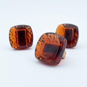 May include: Three square-shaped buttons with rounded corners, featuring a translucent amber-colored resin finish with dark brown mottled patterns and a central rectangular dark brown inlay, each with a gold-toned metal shank on the back.