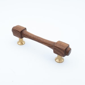 Peut inclure: Une poignée de meuble horizontale composée d'un corps en bois brun foncé avec un centre cylindrique effilé et des extrémités carrées. La poignée est fixée par deux petits supports métalliques de couleur laiton poli orientés vers le bas.