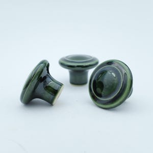 Peut inclure: Trois boutons de meuble en céramique vert foncé avec une finition brillante. Les boutons ont une forme arrondie avec un motif circulaire sur le dessus. Les boutons sont posés sur un fond blanc.