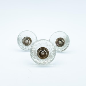 Op de afbeelding: Drie ronde kastknoppen van helder glas met een gestructureerd, radiaal patroon dat vanuit het midden uitstraalt. Elke knop heeft een metalen, zilverkleurige basis met een centraal schroefgat voor montage op meubels.