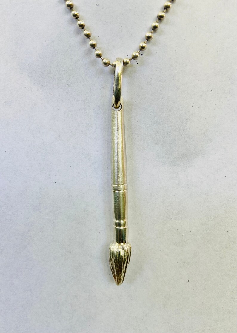 May include: A silver paintbrush pendant necklace. The pendant is shaped like a paintbrush with a detailed brush tip. The necklace is made of a silver chain.