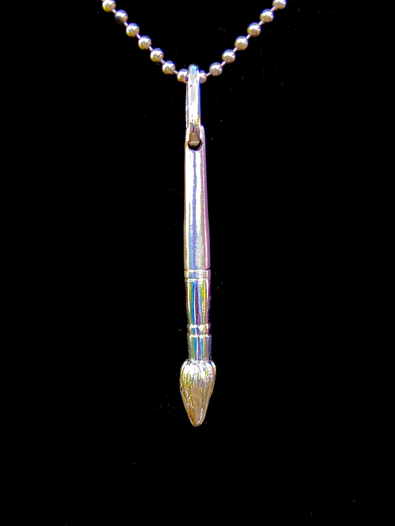 May include: A silver paintbrush pendant necklace. The brush is detailed with a textured bristle tip and a smooth, cylindrical handle.