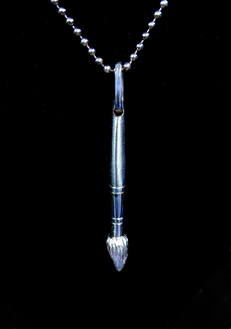 May include: A silver paintbrush pendant necklace with a silver chain. The paintbrush is detailed with a textured brush head.