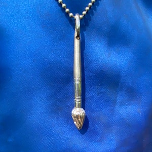 May include: A silver paintbrush pendant necklace on a silver chain. The paintbrush is detailed with a textured brush head.