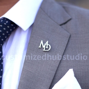 Może przedstawiać: Srebrna przypinka do klapy z inicjałami "MD" przymocowana do szarej marynarki. Przypinka znajduje się na lewej klapie, w pobliżu białej poszetki. Widoczna jest również biała koszula i granatowy krawat w białe grochy.