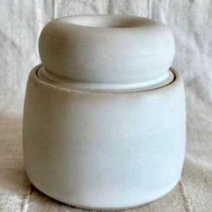 Peut inclure: Un pot en céramique blanc avec un couvercle. Le pot a une base arrondie et un dessus cylindrique avec une ouverture circulaire. Le couvercle repose sur le dessus du pot, créant un aspect lisse et homogène. Le pot est posé sur un fond neutre.