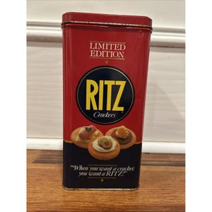 Puede incluir: Una lata rectangular roja de "Edición Limitada" de Ritz Crackers. La lata presenta el logotipo de Ritz en un círculo negro con texto amarillo. La parte inferior de la lata tiene el texto: "When you want a cracker, you want a RITZ."