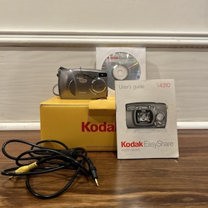 Puede incluir: Una cámara digital Kodak EasyShare plateada, modelo C4310, con una caja amarilla y una guía del usuario. También se incluyen un CD y un cable AV. El logotipo de Kodak es visible en la caja y en la cámara.