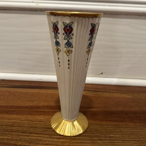 Op de afbeelding: Een crèmekleurige vaas in Art Deco-stijl met een gouden rand en voet. De vaas heeft verticale geribbelde details en is versierd met kleurrijke bloempatronen in rood, blauw en geel. De vaas is ongeveer 18 cm hoog.