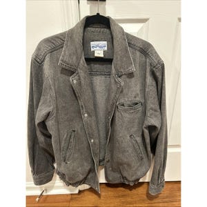 Könnte beinhalten: Graue Jeansjacke mit Knopfleiste, zwei Vordertaschen und einer kleinen Brusttasche. Die Jacke hat einen Kragen und einen gerippten Saum. Die Jacke hängt an einem schwarzen Kleiderbügel.