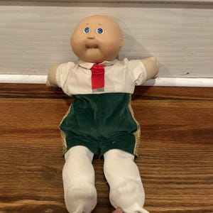 Puede incluir: Una muñeca vintage con cabeza calva y ojos azules. La muñeca lleva una camisa blanca con corbata roja, pantalones cortos de terciopelo verde con ribete dorado y leggings blancos. La muñeca está sobre una superficie de madera.