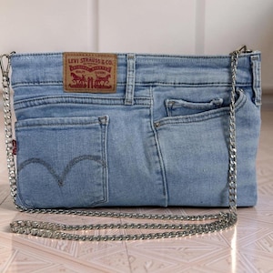 May include: A light blue denim crossbody bag crafted from repurposed jeans, featuring a rectangular shape, a silver-tone metal chain strap, and a tan leather-style brand patch labeled Levi Strauss & Co. with two horses and text.