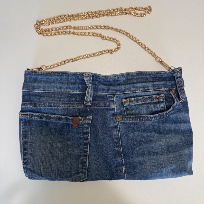 May include: A blue denim handbag constructed from repurposed jeans fabric, featuring a rectangular shape with visible pockets, belt loops, and a gold-tone metal chain strap. The design incorporates classic denim stitching and metal rivets.