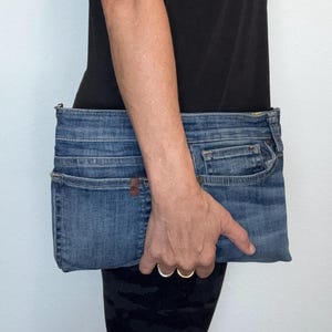 May include: A rectangular clutch bag crafted from blue denim fabric, featuring the distinct pockets, seams, and waistband details of a pair of jeans. The design includes a top zipper closure and maintains the authentic texture and wash of denim material.