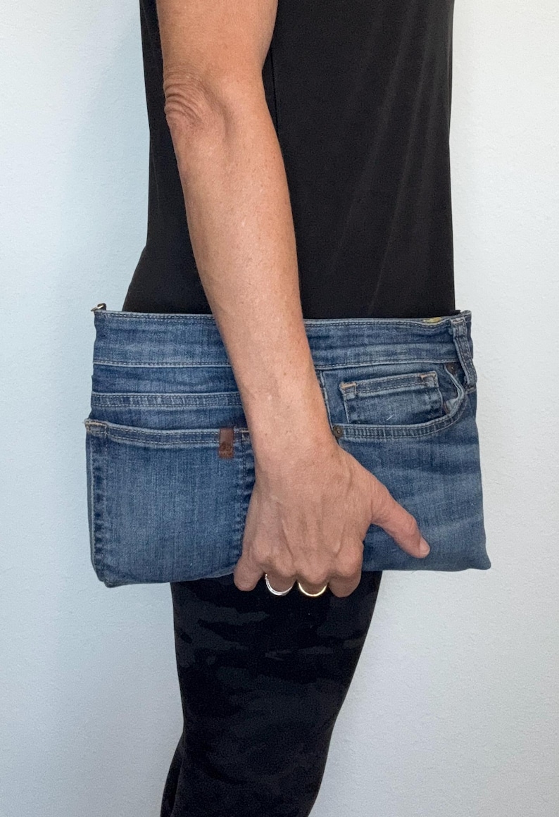 May include: A rectangular clutch bag crafted from blue denim fabric, featuring the distinct pockets, seams, and waistband details of a pair of jeans. The design includes a top zipper closure and maintains the authentic texture and wash of denim material.