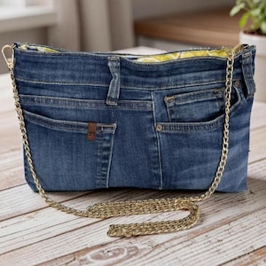 May include: A blue denim handbag constructed from repurposed jeans, featuring visible pockets, belt loops, and a metal button. The bag includes a gold-tone metal chain shoulder strap and a bright yellow interior lining with a white lemon pattern.