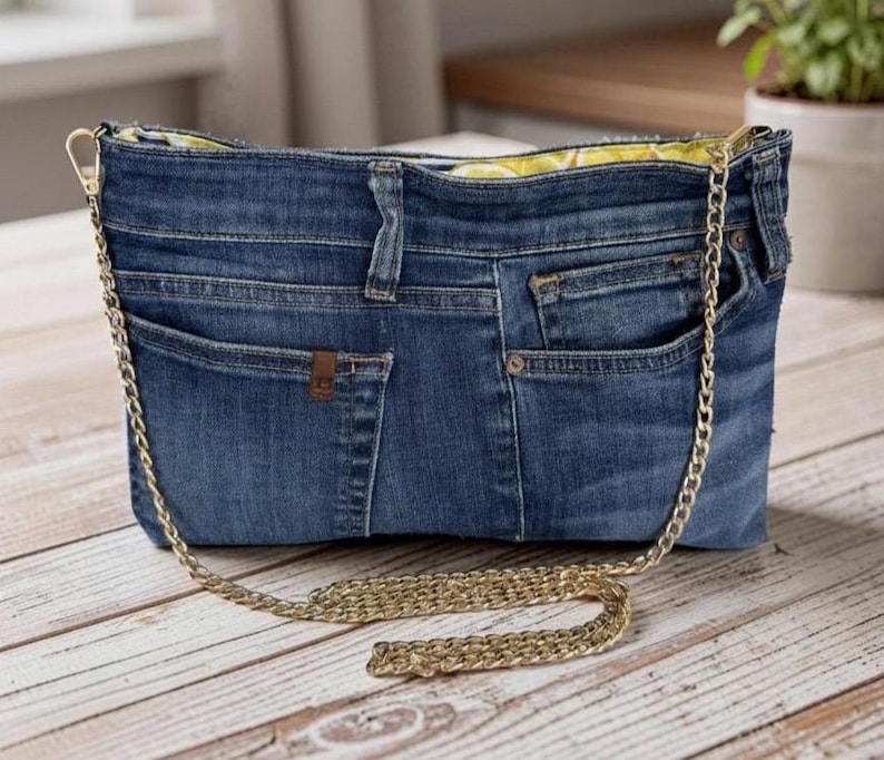 May include: A blue denim handbag constructed from repurposed jeans, featuring visible pockets, belt loops, and a metal button. The bag includes a gold-tone metal chain shoulder strap and a bright yellow interior lining with a white lemon pattern.