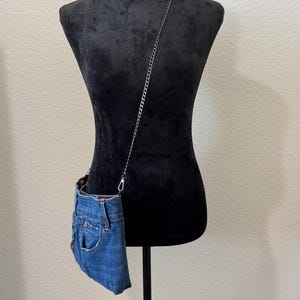 May include: A crossbody bag crafted from blue denim fabric, featuring a visible pocket and waistband detail. The bag is attached to a thin, dark metal chain strap that hangs diagonally across the torso, showcasing a casual and functional design.