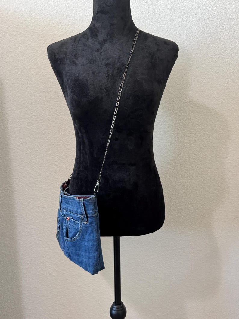 May include: A crossbody bag crafted from blue denim fabric, featuring a visible pocket and waistband detail. The bag is attached to a thin, dark metal chain strap that hangs diagonally across the torso, showcasing a casual and functional design.