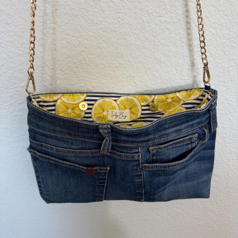 May include: A blue denim handbag featuring a gold chain strap and a snap closure. The interior lining is decorated with a yellow lemon pattern on a navy and white striped background. A small white label with the text Indy Bags is visible inside.