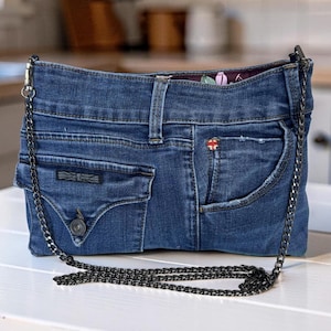 May include: A blue denim handbag constructed from repurposed jeans, featuring a front pocket with a button closure, a small red and white logo tag, and a dark metal chain shoulder strap. The interior lining displays a floral pattern.