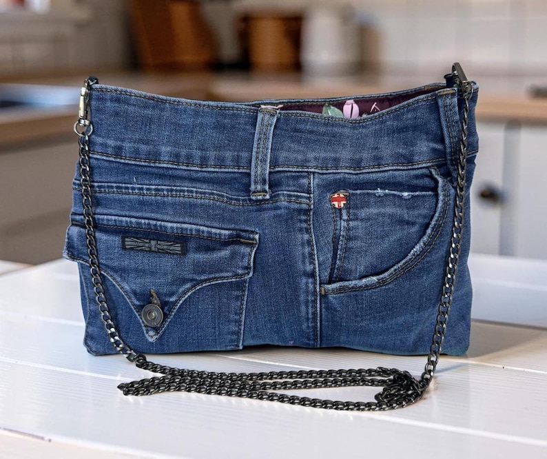 May include: A blue denim handbag constructed from repurposed jeans, featuring a front pocket with a button closure, a small red and white logo tag, and a dark metal chain shoulder strap. The interior lining displays a floral pattern.