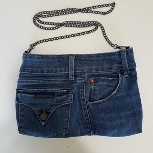 May include: A blue denim handbag crafted from repurposed jeans material, featuring a dark metal chain strap, a front flap pocket with a button closure, and a side pocket. The bag displays a small red and white flag logo on the upper right side.