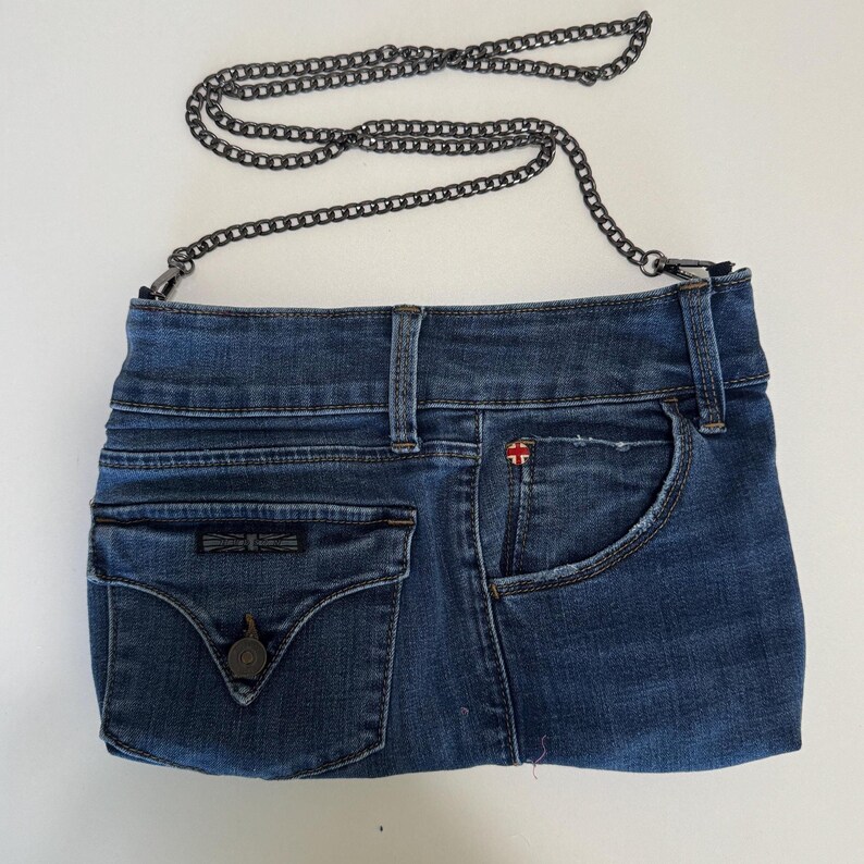 May include: A blue denim handbag crafted from repurposed jeans material, featuring a dark metal chain strap, a front flap pocket with a button closure, and a side pocket. The bag displays a small red and white flag logo on the upper right side.