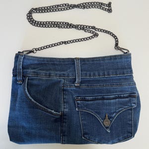 May include: A blue denim handbag constructed from repurposed jeans fabric, featuring a waistband top, a side pocket, and a flap pocket with a metal button closure. The bag includes a detachable dark gray metal chain strap with lobster clasps.