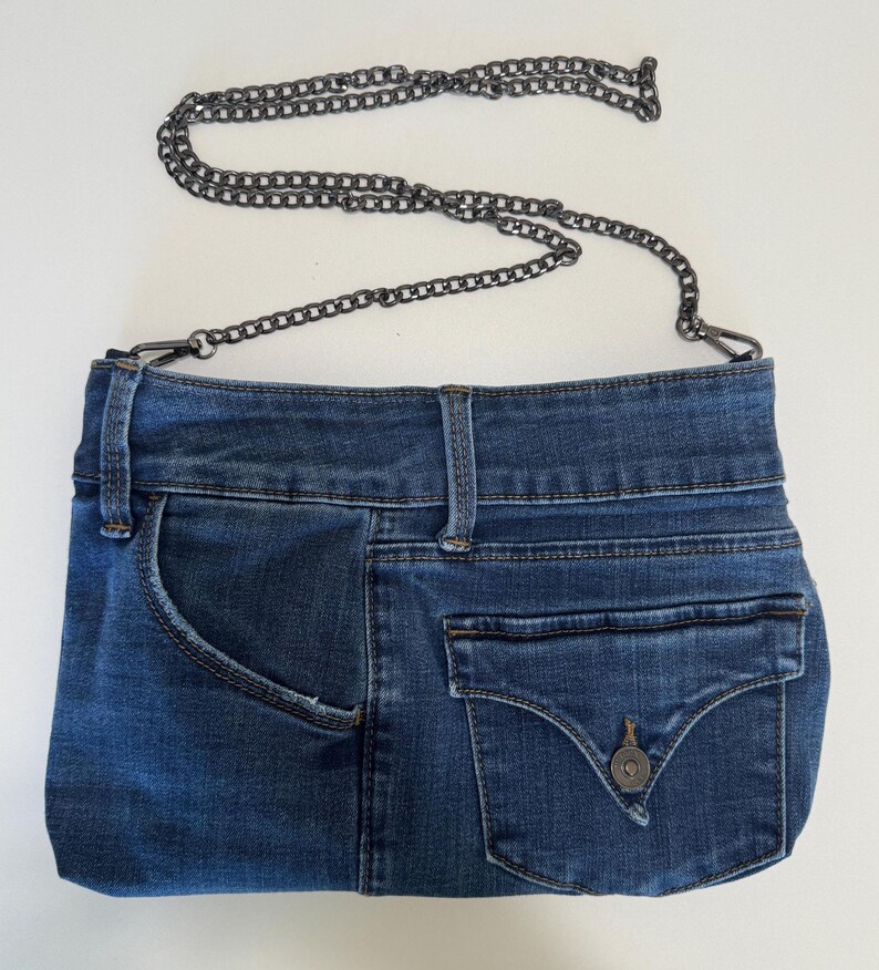 May include: A blue denim handbag constructed from repurposed jeans fabric, featuring a waistband top, a side pocket, and a flap pocket with a metal button closure. The bag includes a detachable dark gray metal chain strap with lobster clasps.