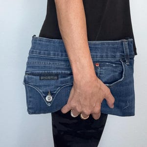 May include: A blue denim clutch bag constructed from repurposed jeans fabric, featuring a visible waistband, a front pocket with a metal button closure, and a small red logo tag near the pocket edge, showcasing authentic denim stitching and texture.
