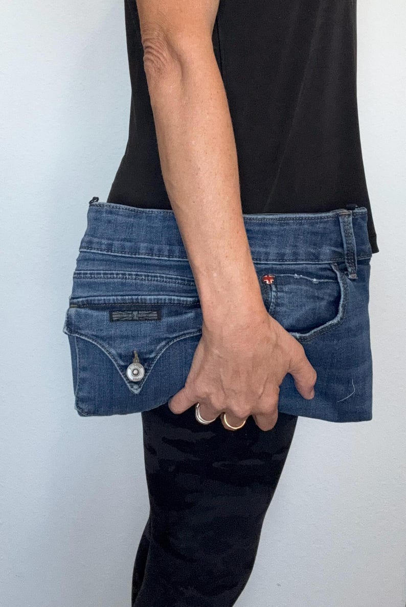 May include: A blue denim clutch bag constructed from repurposed jeans fabric, featuring a visible waistband, a front pocket with a metal button closure, and a small red logo tag near the pocket edge, showcasing authentic denim stitching and texture.
