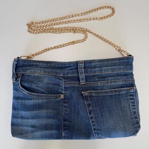 May include: A rectangular crossbody bag crafted from blue denim fabric, featuring a front pocket, a waistband detail with belt loops, and a back pocket. The bag includes a detachable gold-tone metal chain strap attached with lobster claw clasps.