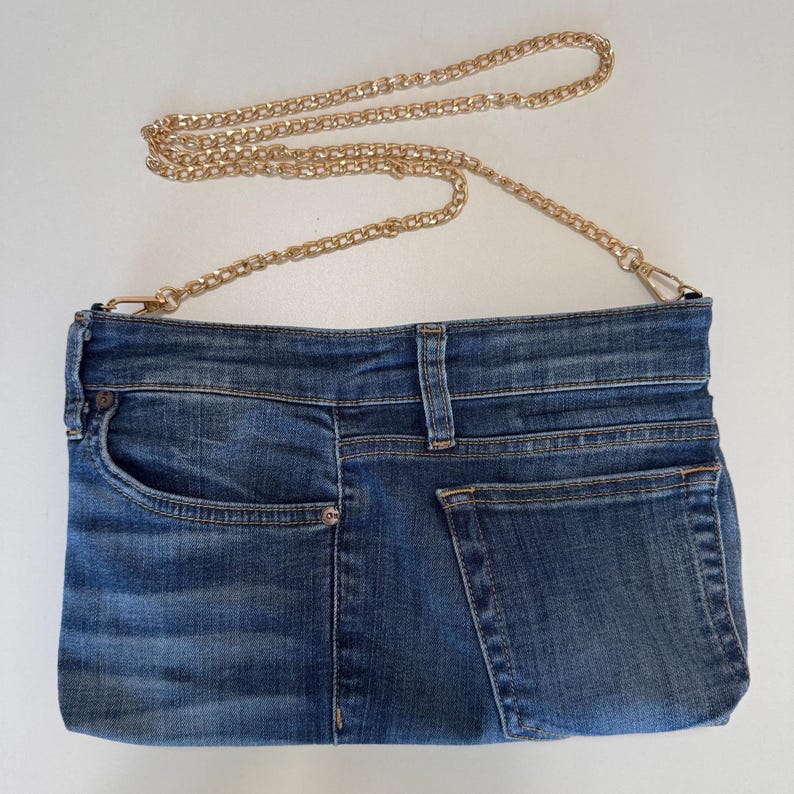 May include: A rectangular crossbody bag crafted from blue denim fabric, featuring a front pocket, a waistband detail with belt loops, and a back pocket. The bag includes a detachable gold-tone metal chain strap attached with lobster claw clasps.