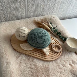 Op de afbeelding: Een handgemaakt gehaakt schildpad speelgoed in groen en crème, rustend op een houten dienblad met een houten ring bijtring. De schildpad heeft een ronde kop en schild, met crèmekleurige vinnen. De achtergrond omvat een zacht tapijt en decoratieve elementen.