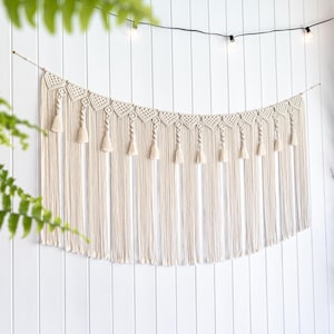 Könnte beinhalten: Ein cremefarbener Makramee-Wandbehang mit geschwungenem Design. Die Oberseite hat ein geometrisches Muster mit Quasten darunter. Er hängt an einer weißen Holzwand, darüber Lichterketten. Ungefähr 100 cm breit.