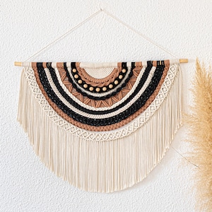Könnte beinhalten: Ein halbkreisförmiger Makramee-Wandbehang mit einem Holzdübel. Das Design zeigt cremefarbene, schwarze und hellbraune Bänder, Holzperlen und Fransen. Dieses dekorative Stück verleiht jedem Raum einen Hauch von Bohème.