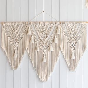 May include: Cream-colored macrame wall hanging with geometric patterns and tassels. The handcrafted textile art piece is suspended from a wooden dowel, adding a bohemian touch to home decor. The wall hanging is approximately 40 inches wide.