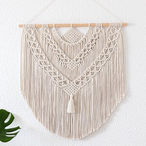Könnte beinhalten: Cremefarbener Makramee-Wandbehang mit Holzstab. Das Design zeigt aufwendige Knotenmuster und eine Quaste am unteren Ende. Die Wanddekoration im Boho-Stil verleiht jedem Raum eine besondere Note und Textur.