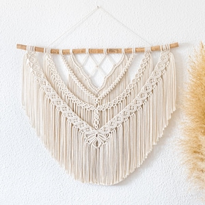 Peut inclure: Suspension murale en macramé crème avec un motif géométrique, suspendue à une cheville en bois naturel. Le nouage complexe crée une esthétique bohème texturée. La décoration murale présente un motif symétrique et de longues franges.