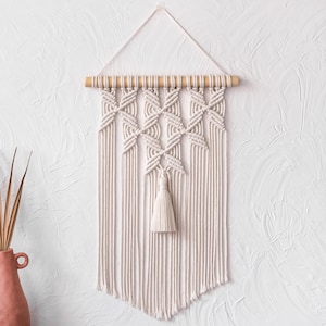 Pode incluir: Uma tapeçaria de parede em macramé creme com um pino de madeira e uma borla. O design apresenta padrões geométricos e fios verticais, criando uma estética boêmia texturizada. A decoração da parede é exibida contra uma parede branca texturizada.