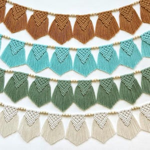 Op de afbeelding: Een close-up van verschillende macramé slingers in tinten bruin, blauwgroen, groen en crème. Elke slinger heeft gefranjerde, geometrische vormen geregen op een houten kralenketting. De achtergrond is een effen wit oppervlak.