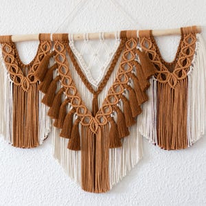 May include: A macrame wall hanging with a wooden dowel and detailed knotwork. The design uses brown, cream, and white threads, with tassels and geometric patterns. This handmade piece is a stylish addition, bringing a bohemian feel to a room.