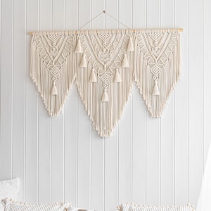 May include: Cream-colored macrame wall hanging with a geometric design and tassels. The wall decor is mounted on a wooden dowel and hangs against a white paneled wall. The macrame features a central design flanked by two symmetrical panels.