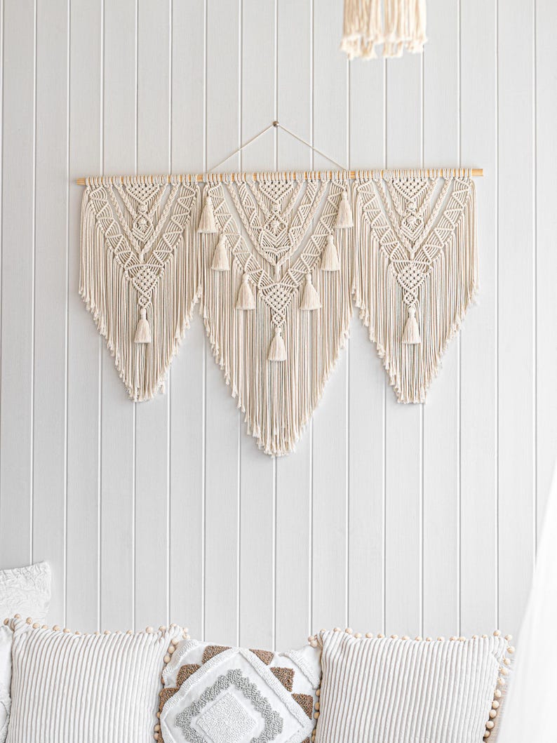 May include: Cream-colored macrame wall hanging with a geometric design and tassels. The wall decor is mounted on a wooden dowel and hangs against a white paneled wall. The macrame features a central design flanked by two symmetrical panels.